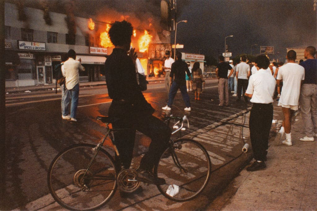 A view of businesses beginning to burn on Pico Boulevard near Hayworth Avenue, onlookers gathering, a young man dressed in black pausing on bicycle watching fire during the Rodney King Riots, and the sky black with smoke in daylight on April 30, 1992 in Los Angeles, California. (Photo by Lindsay Brice/Getty Images)